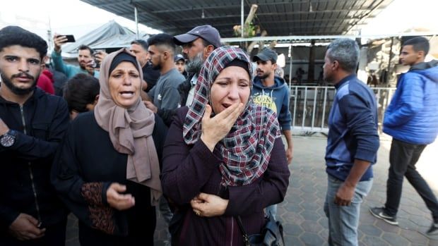 Two women wearing headcoverings are prominent in a photo, standing outside with pained expressions. Several others, mostly men, are seen in the background.