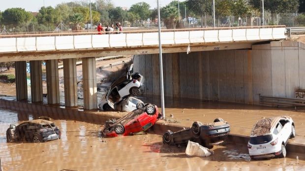 Two overturned cars and three other vehicles are shown in muddy water by a bridge.