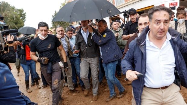 The king of Spain walks under an umbrella while people around him are pelted with mud.