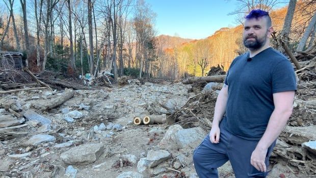 A person looks at the camera while standing amid debris and fallen branches.