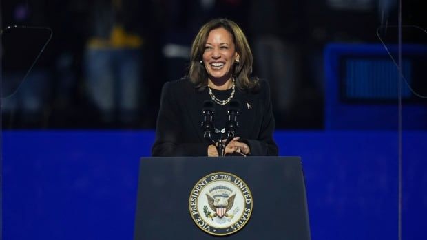 Kamala Harris, wearing a dark blazer and her signature necklace, speaks at a podium in front of a large crowd of people holding political signs. 