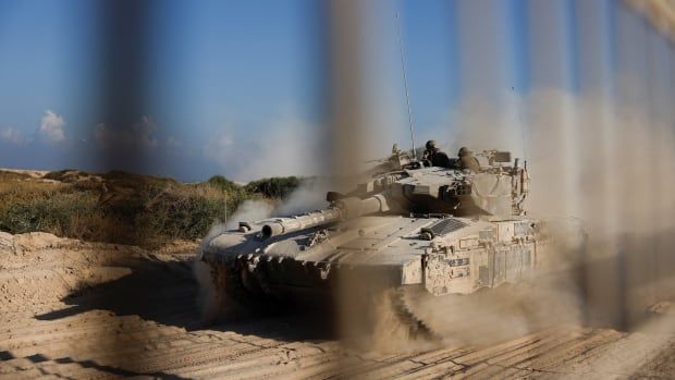 A tank drives down a dusty path, visible through the blurred bars of some sort of fence. 