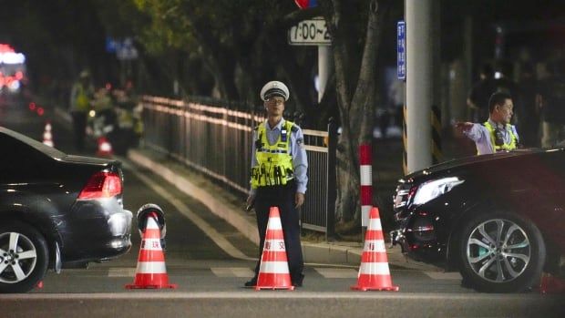 An Asian man wearing a glasses and a police or security type uniform stands near a roadside with a couple of pylons laid down in a nighttime photo.