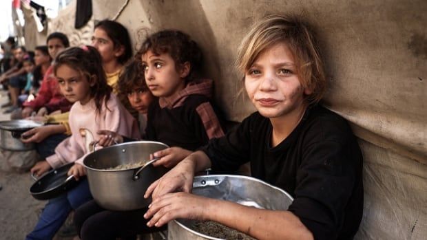 Children sit on a bench in a tent holding tin pots.