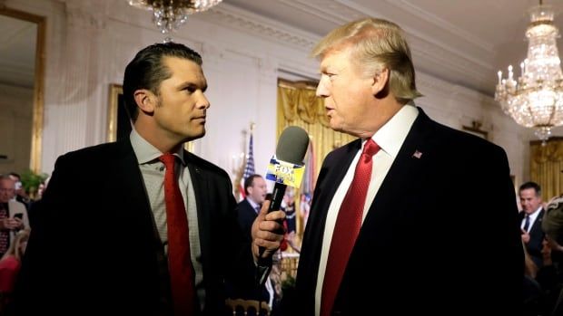 A dark haired, clean shaven man in a suit and tie holds a microphone near the face of an older, clean shaven light-haired man in a suit and tie who is speaking.