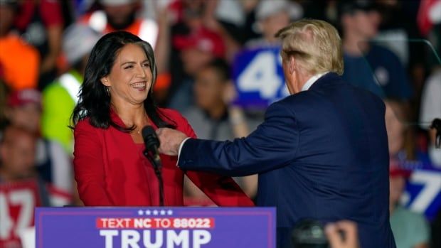 A dark-haired woman wearing a red suit jacket smiles from behind a podium as a man in a suit and tie approaches with his arm out.