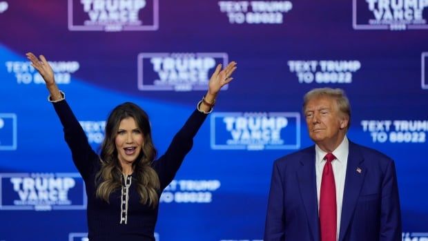 A dark haired woman raises her arms above her head while an older clean shaven man in a suit and tie stands nearby. Both are on a stage.