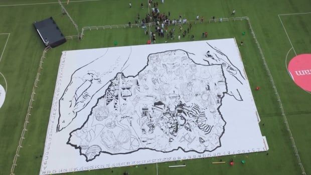 Aerial view of a massive drawing laid out over several canvasses encompassing the bilk of a football field, with people gathered around its edges. The drawing shows two large hands encircling a map of Nigeria, filled with images of people, masks, relics, foods, and more.