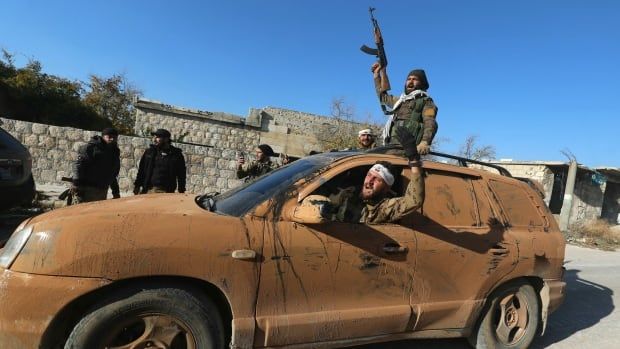 People in military garb lean out of a moving car to cheer and wave weapons.