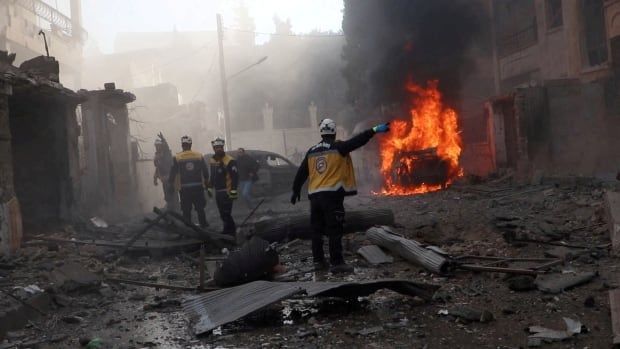 Men in white helmets walk on a street filled with rubble. In the background, a car is in flames.