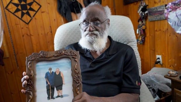 A solemn man with a white beard holds a framed photo of his wife with him on a beach vacation.