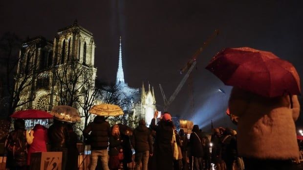 Crowds with umbrellas are seen in the foreground as a cathedral surrounded by construction cranes is seen in the background.