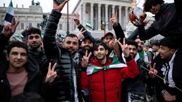 Dozens of smiling Syrians stand outside flashing peace signs. Some are wrapped in their country's green, white and red flag, and some are holding musical instruments.