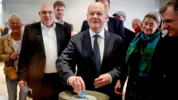 A bald man in a suit and tie smiles while dropping a piece of paper into box. He is standing, surrounded by men and women. 