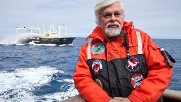 Man standing in a bright orange jacket. A whaling vessel is on the water in the distance behind him.