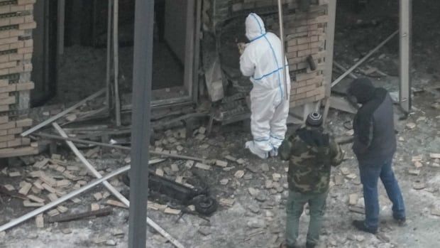 A man in a white hazmat suit inspects the wall of a burned brick building. Behind him, two other people look on amid rubble.