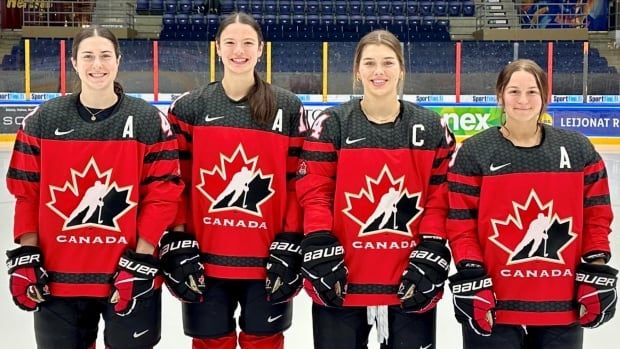 Four women in Hockey Canada jerseys, each with an A or a C on the sweater, pose on the ice.