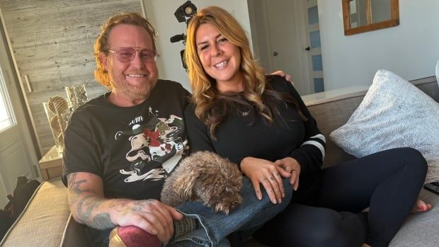 Man and woman sit on a couch smilling. A small brown dog sits in the mans lap asleep. 