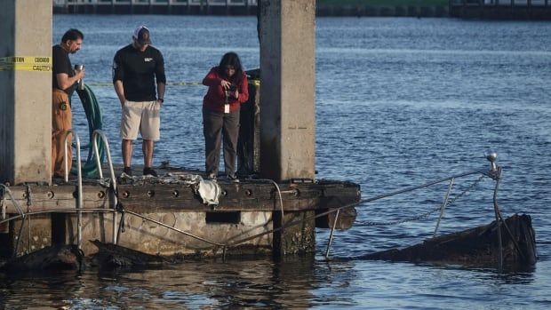 People standing on the edge of a pier survey the charred remains of a boat in the water.