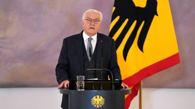 A man wearing a suit and glasses stands at a podium with a red, yellow and black flag behind him.