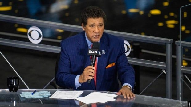 A male sportscaster wearing a suit and tie speaks into a microphone while sitting at a desk.