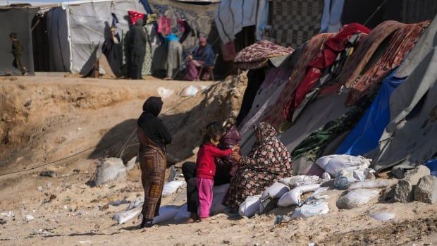 A little girl hands another person something in the middle of the image, while a third person stands behind her. The person being handed something is sitting on a slope with a tent made out of various cloths behind them. In the distance, more tent structures and people can be seen. 