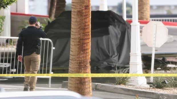 A person behind police tape holds a cellphone to their ear as they walk toward a vehicle covered in a tarp.