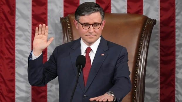 A man in a suit is at a desk or podium, holding his right hand up in the air and with his left hand placed flat on top of a book on the desk. 