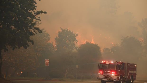 A fire truck drives on a street blanketed in smoke as a fire burns behind the treeline.