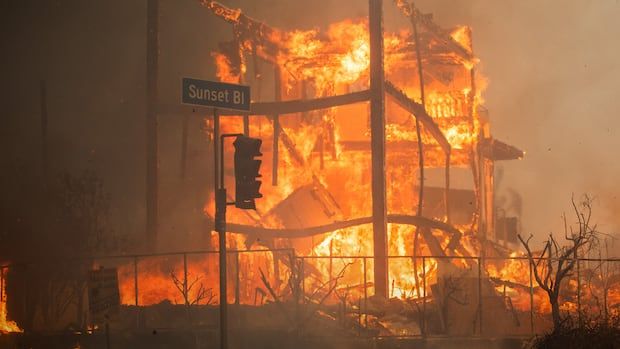 A street signs in front of a building engulfed by flames. 