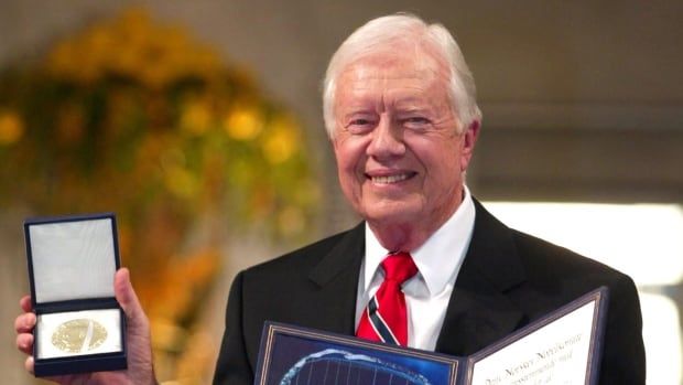 Wearing a dark suit, Jimmy Carter smiles while holding his Nobel Peace Prize.