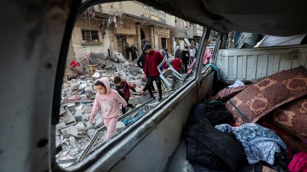 People are framed through the window of a car carrying belongings and walking amid rubble.