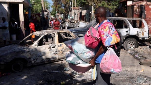 A many carries many bags among burned-out vehicles on the street.