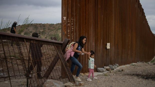 A mother and her daughter enter the US next to a rusty steel or iron fence in the Sonoran desert