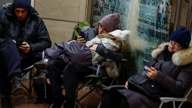 A woman in glasses and winter hat and clothing is seated, holding a small child in winter clothing while looking at her phone. On either side of the woman and child are people also in winter clothing seated on chairs looking at their devices.