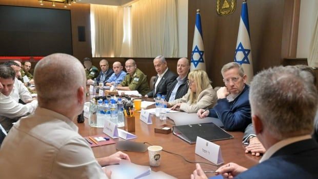 Several people, men and women, some in suit and business attire and others in military uniforms, are shown seated at a long table in front of Israel flags.