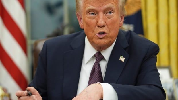 A man with orange-grey hair, wearing a navy suit, holds a pen as he signs documents at a desk, in front of an American flag.