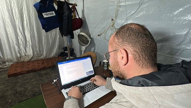 Diaa Al-Ustaz works on a laptop while his press flak jacket hangs on a coat stand in the background.