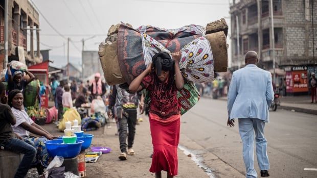 A person carries a mattress and other belongings on their back as they walk on a busy street.