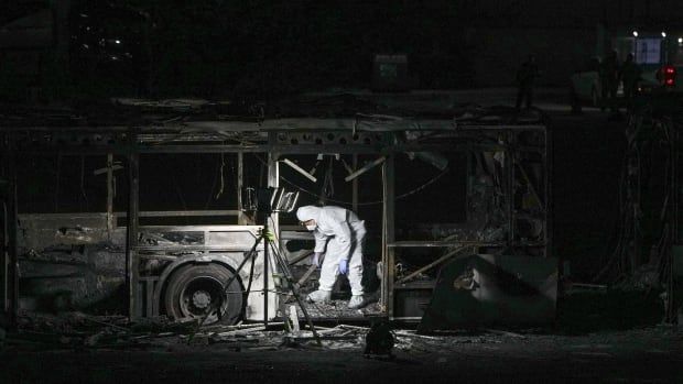 A police officer in a white hazmat-like suit inspects the burned wreckage of a bus at night.