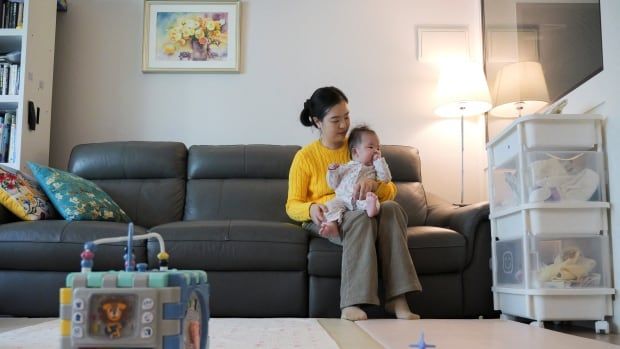 An East Asian woman wearing a yellow sweater sits on a black leather couch holding an infant child.