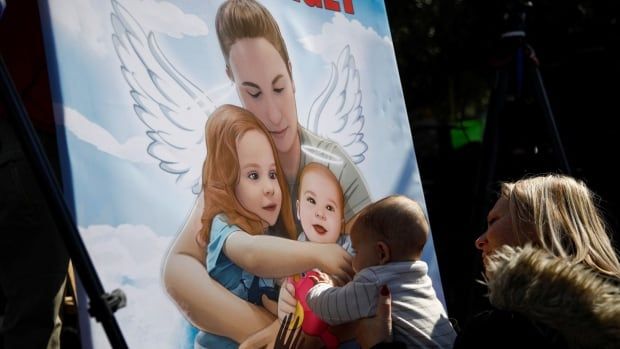 A woman holds up a baby to a poster of an image of a woman with two small children. The woman on the poster has angel wings.