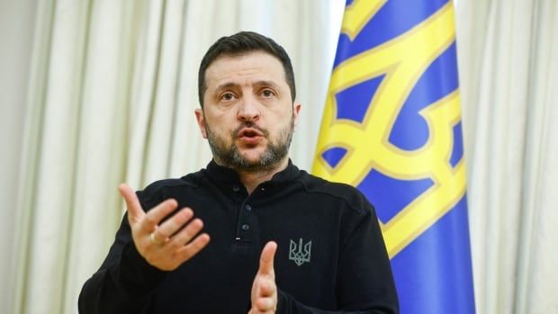 A bearded man in dark clothing gestures with his hand while speaking in front of a blue and yellow flag.