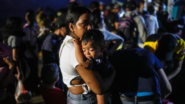 A brown-skinned woman wearing a tshirt and jeans holds a small boy in her arms. It's a nighttime scene, and dozens of people are behind her.