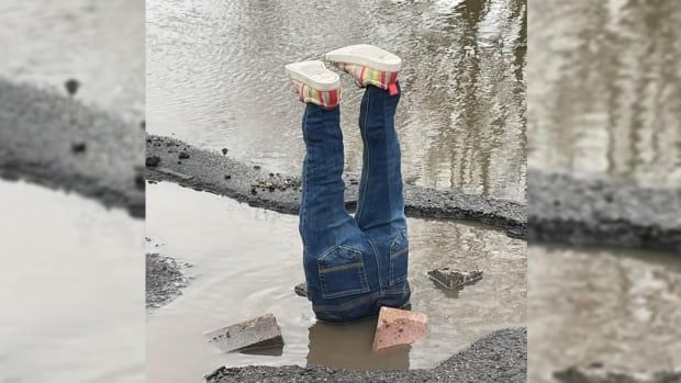 A pair of fake legs in jeans and sneakers sticks straight out of a puddle-and-brick-filled pothole on a gray, rainy day.