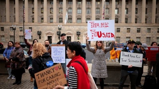 Several people are shown on a sidewalk holding signs outside a building in an apparent demonstration.