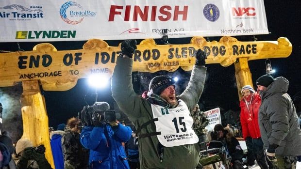 Male dog musher, raises arms in air to celebrate win with finish line sign in the background.