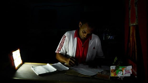 A person uses a lamp for light as they write at a table during a blackout.