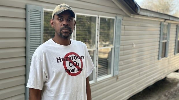 An adult Black man stands outside wearing a white t-shirt with the words "hazardous CO2 pipeline" with a no-smoking cross-out symbol on top.