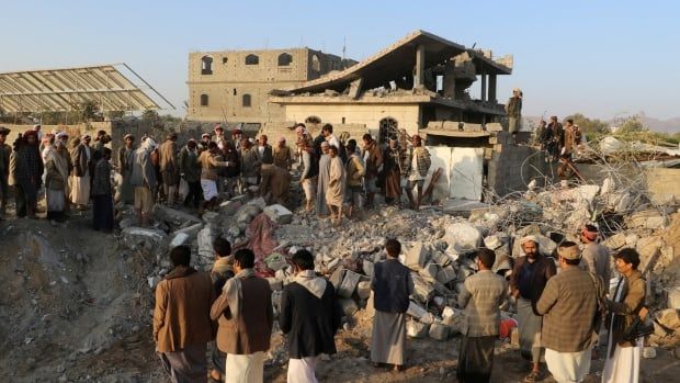 People gather around a crater strewn with debris from destroyed buildings.